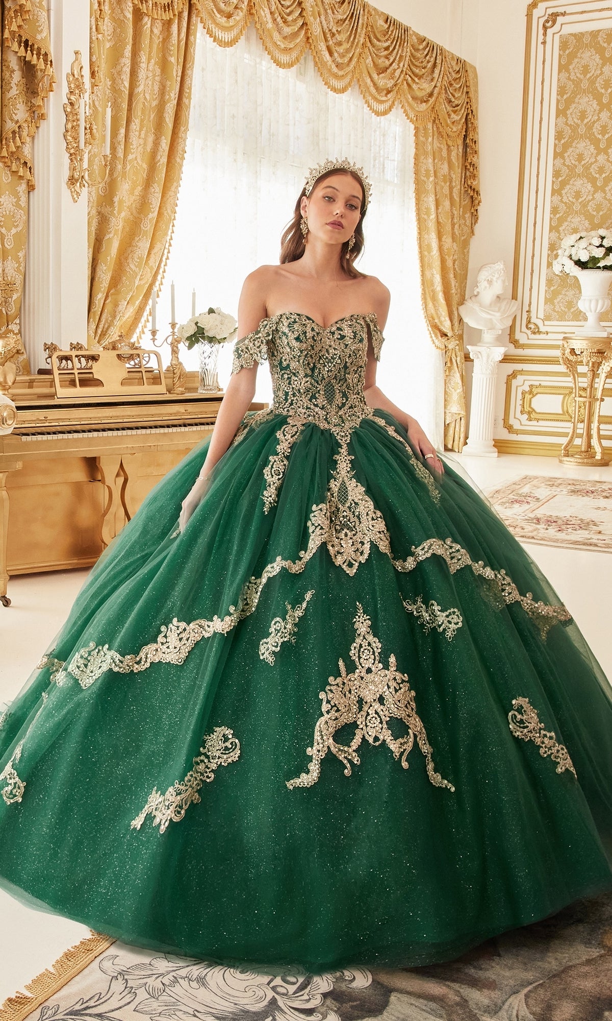 A woman stands in an opulent room with gold furnishings and drapes wearing the Ladivine 15711 Quinceañera Dress, a green ball gown adorned with gold embroidery and glitter tulle.